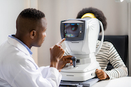 Brown skinned man or doctor interpreting results on a vision machine, while brown skinned woman looks into the machine.